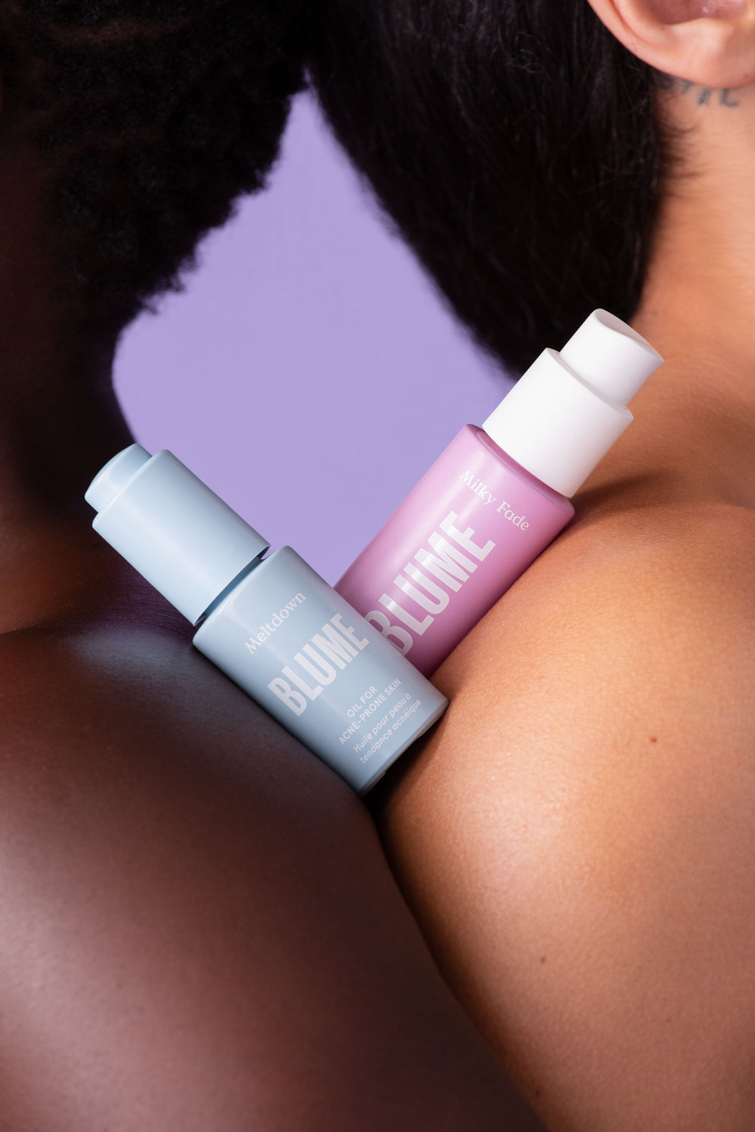 Blume Meltdown Acne Oil and Milky Fade Serum resting on two diverse shoulders against a lavender background, highlighting inclusive, gentle skincare