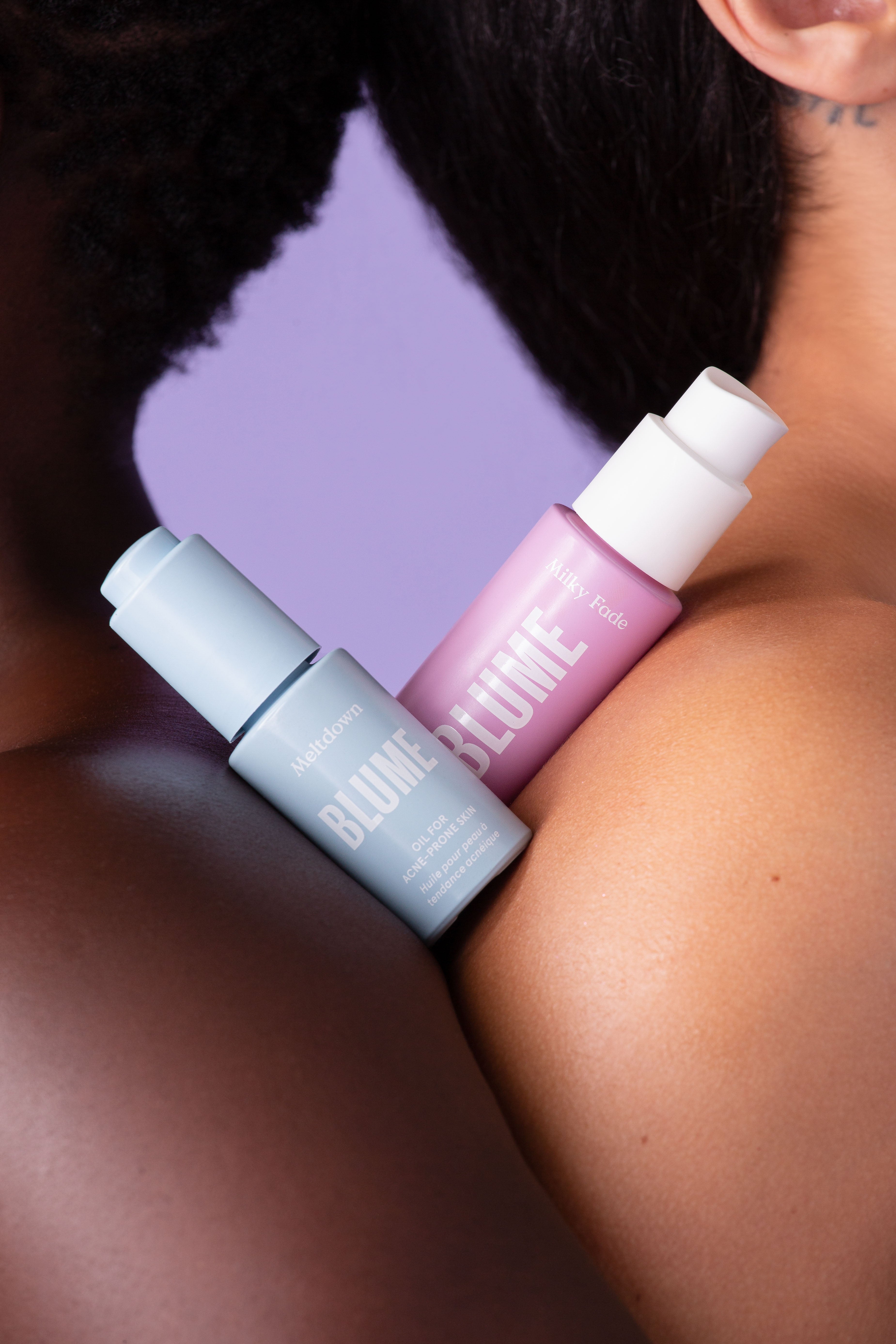 Blume Meltdown Acne Oil and Milky Fade Serum resting on two diverse shoulders against a lavender background, highlighting inclusive, gentle skincare