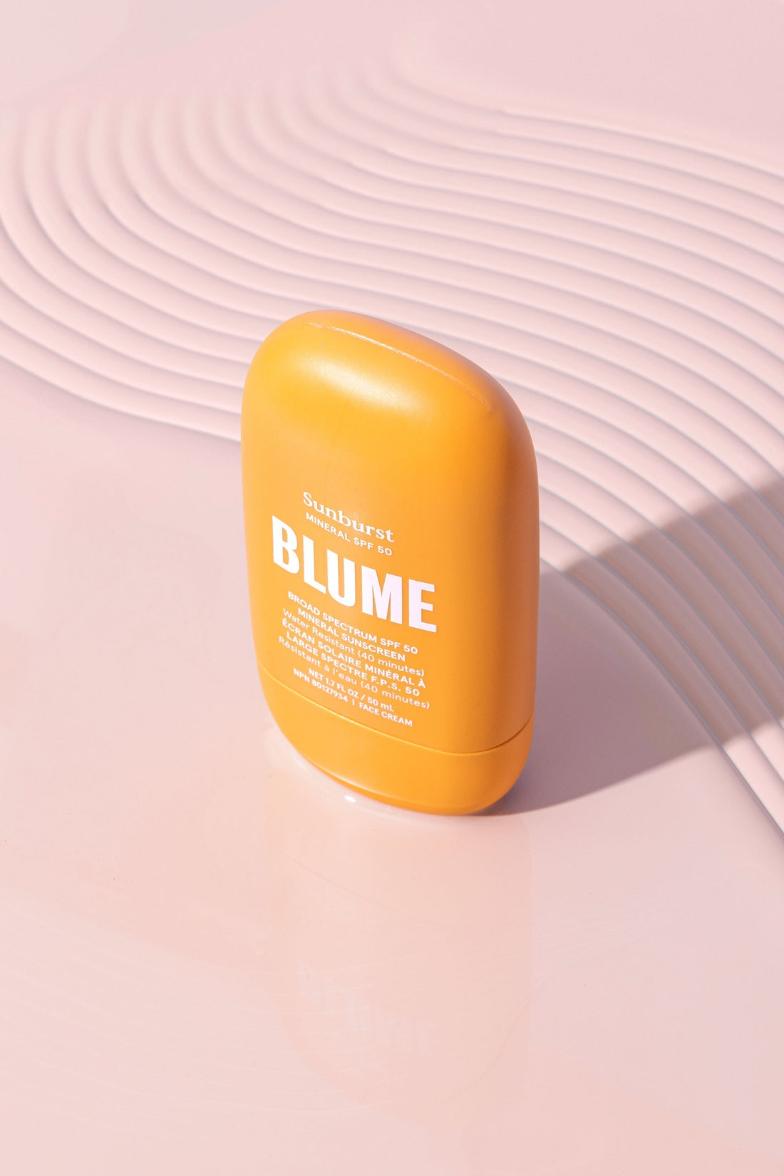 Blume Sunburst Mineral SPF 50 sunscreen standing upright on a glossy pink surface with curved ripple textures and soft lighting.