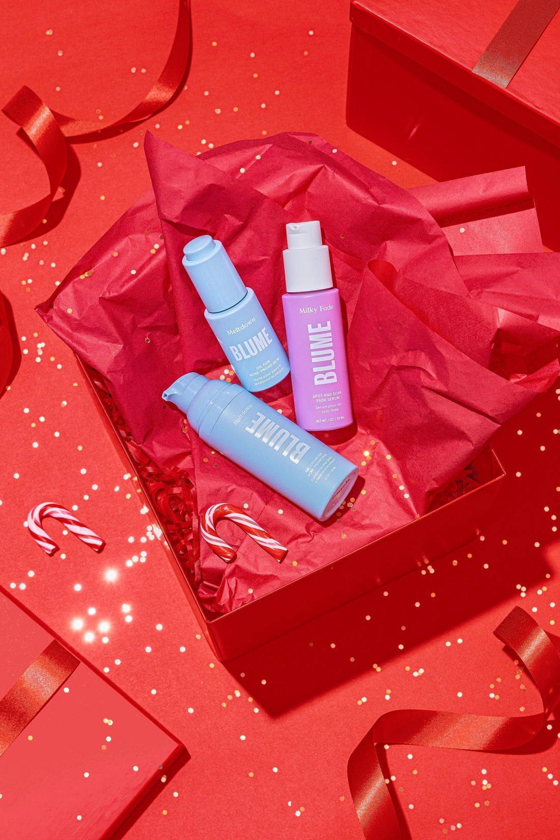 Blume skincare holiday gift set with Milky Fade serum and Meltdown acne products in a red gift box with festive tissue paper, candy canes, and ribbon on a red background.