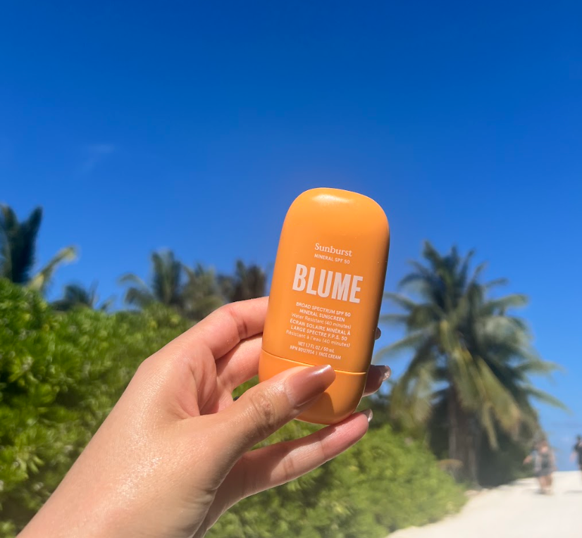 Hand holding Blume Sunburst Mineral SPF 50 sunscreen with a tropical beach and palm trees in the background, perfect for reef-safe sun protection