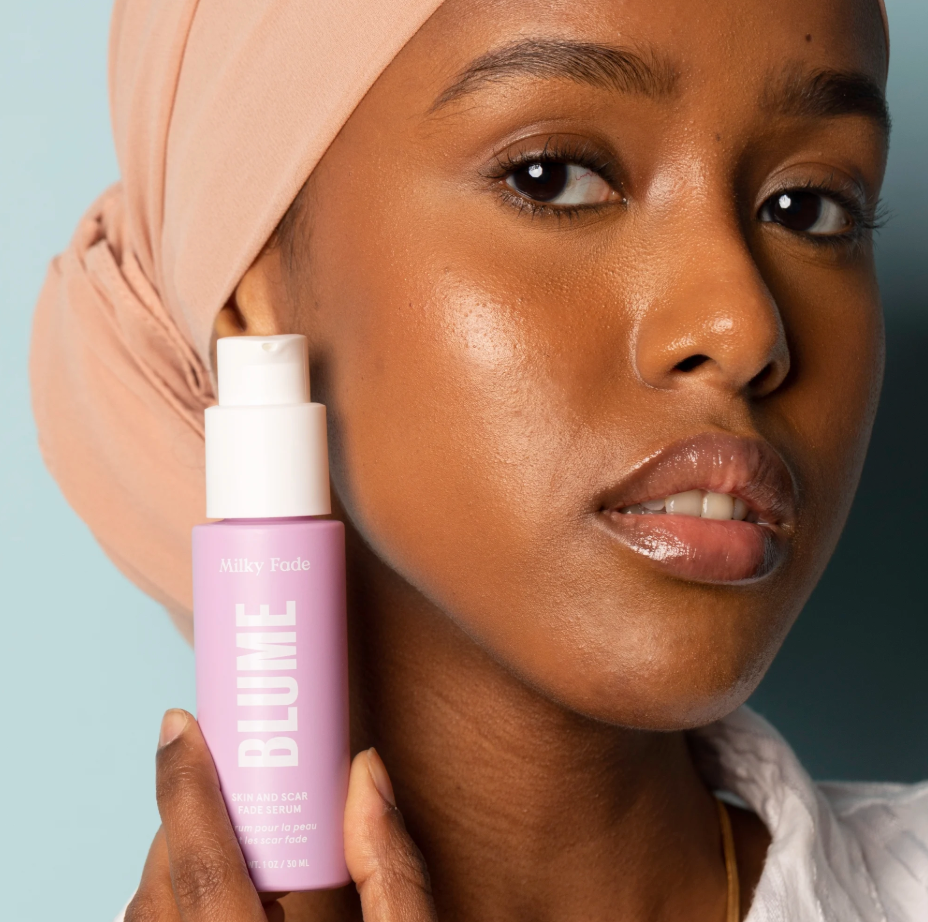 Woman with radiant skin holding Blume Milky Fade skin and scar fade serum against light blue background
