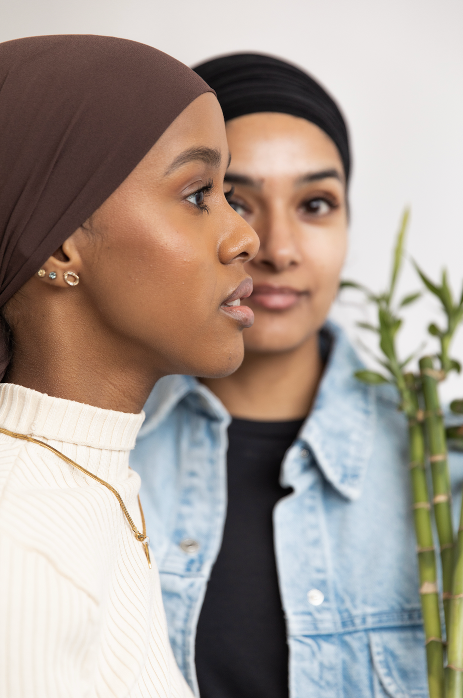 One woman wearing a cream ribbed turtleneck and gold jewelry, the other wearing a denim jacket holding bamboo, representing natural skincare and mindful self-care