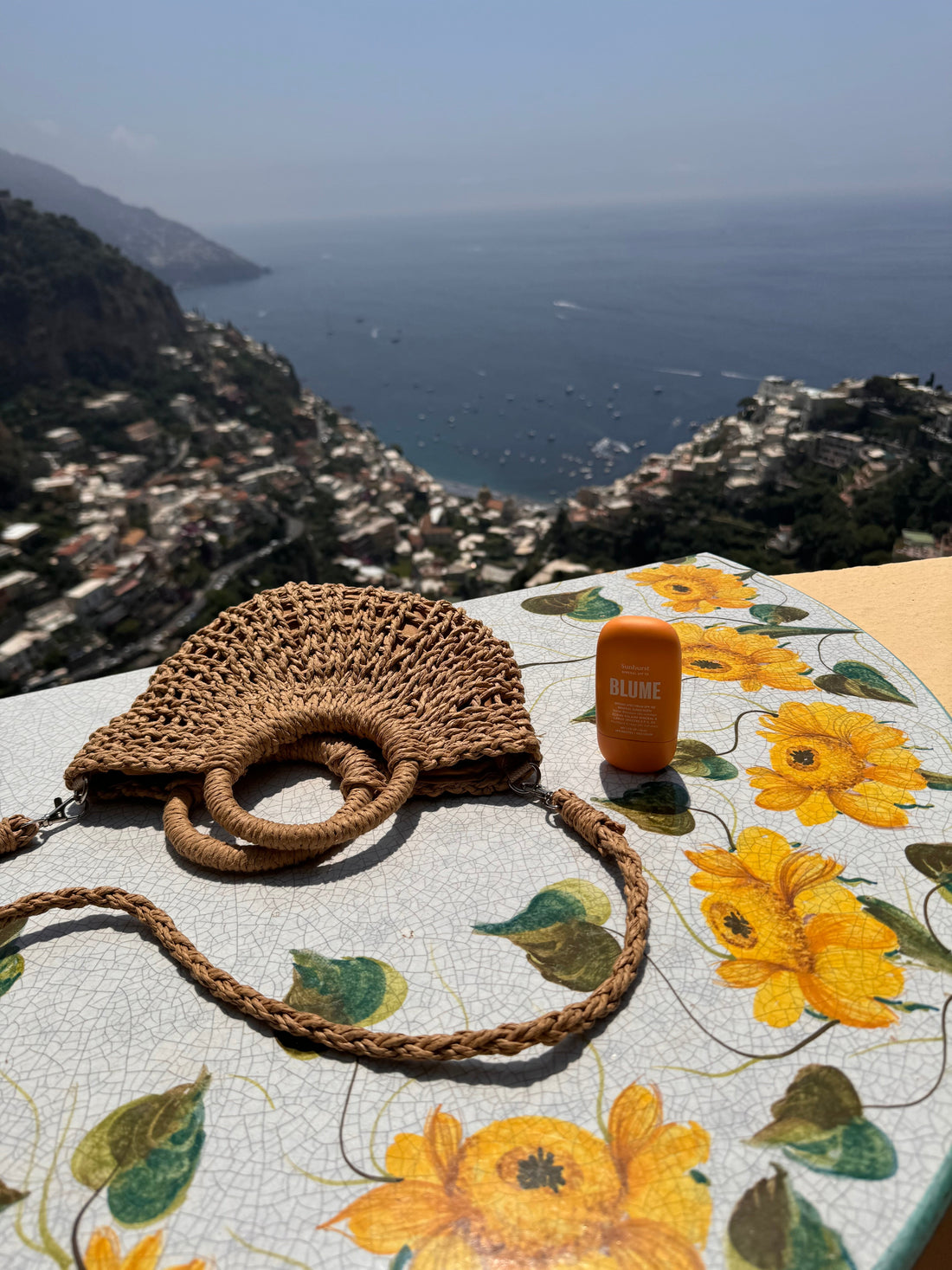 Blume Sunburst SPF 50 mineral sunscreen on a sunflower-painted table with a woven summer bag and a scenic ocean view in Positano, Italy; perfect for travel-friendly sun protection.
